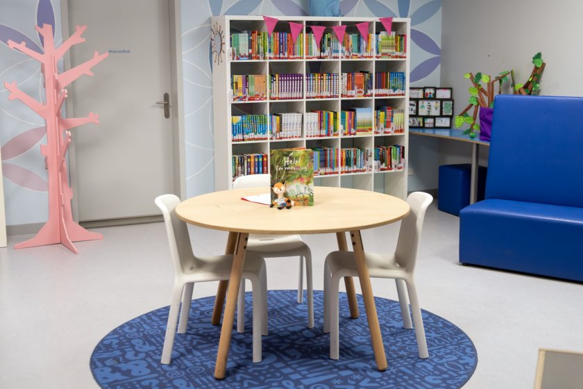 Een foto van een deel van de schoolbibliotheek. Op de foto zie je een ronde tafel, met twee stoelen en een boekenkast vol met boeken.